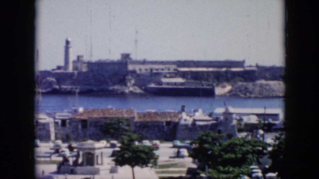 1956 Cuba 8mm frame showing the fortress and lighthouse across the harbor, framed by low buildings, trees, cars, and a small open civic foreground.