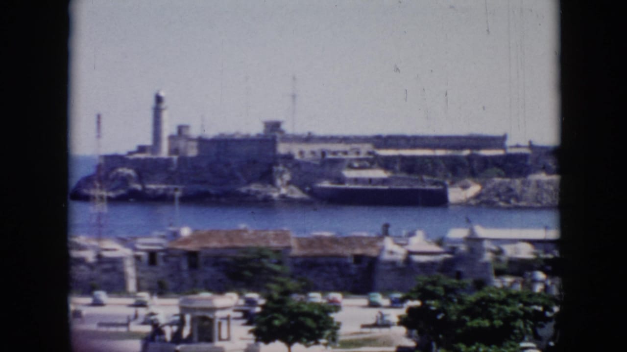 1956 Cuba 8mm frame with a soft, slightly blurred view of a coastal fortress and lighthouse across the water, with waterfront structures and trees below.