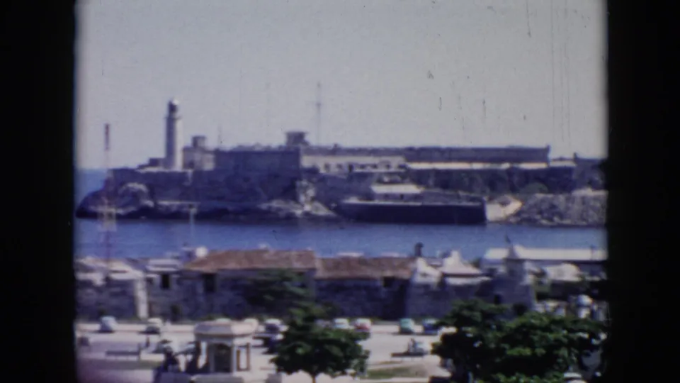 1956 Cuba 8mm frame with a soft, slightly blurred view of a coastal fortress and lighthouse across the water, with waterfront structures and trees below.