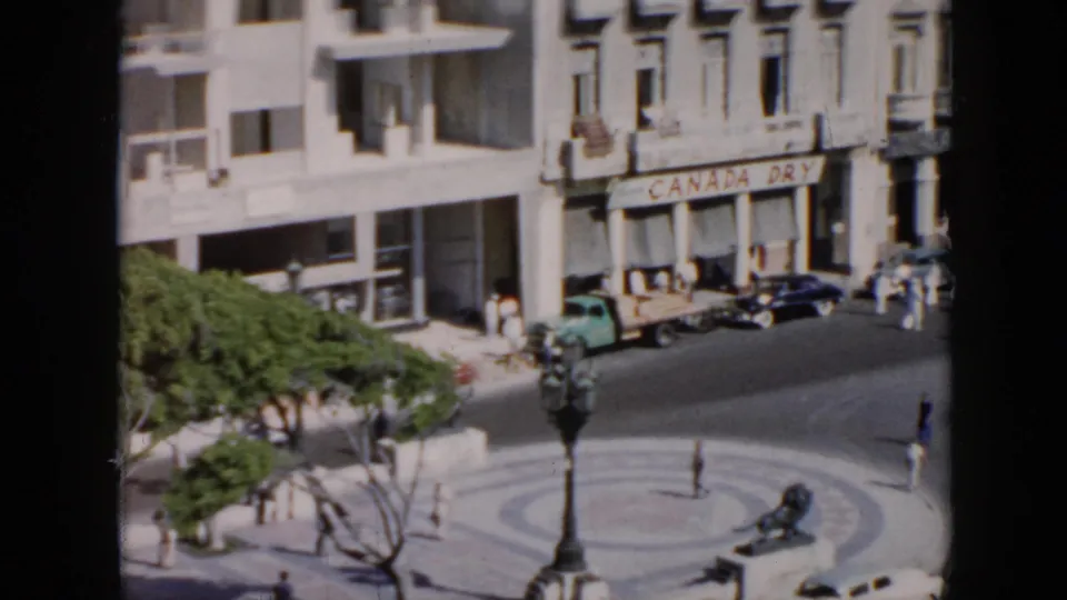 Elevated 1950s Cuba street view showing a circular plaza, pedestrians, vintage vehicles, trees, and storefront buildings.