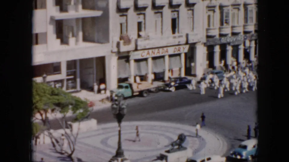1950s Cuba street view with white-uniformed marchers passing near a circular plaza, cars, pedestrians, and a Canada Dry sign.