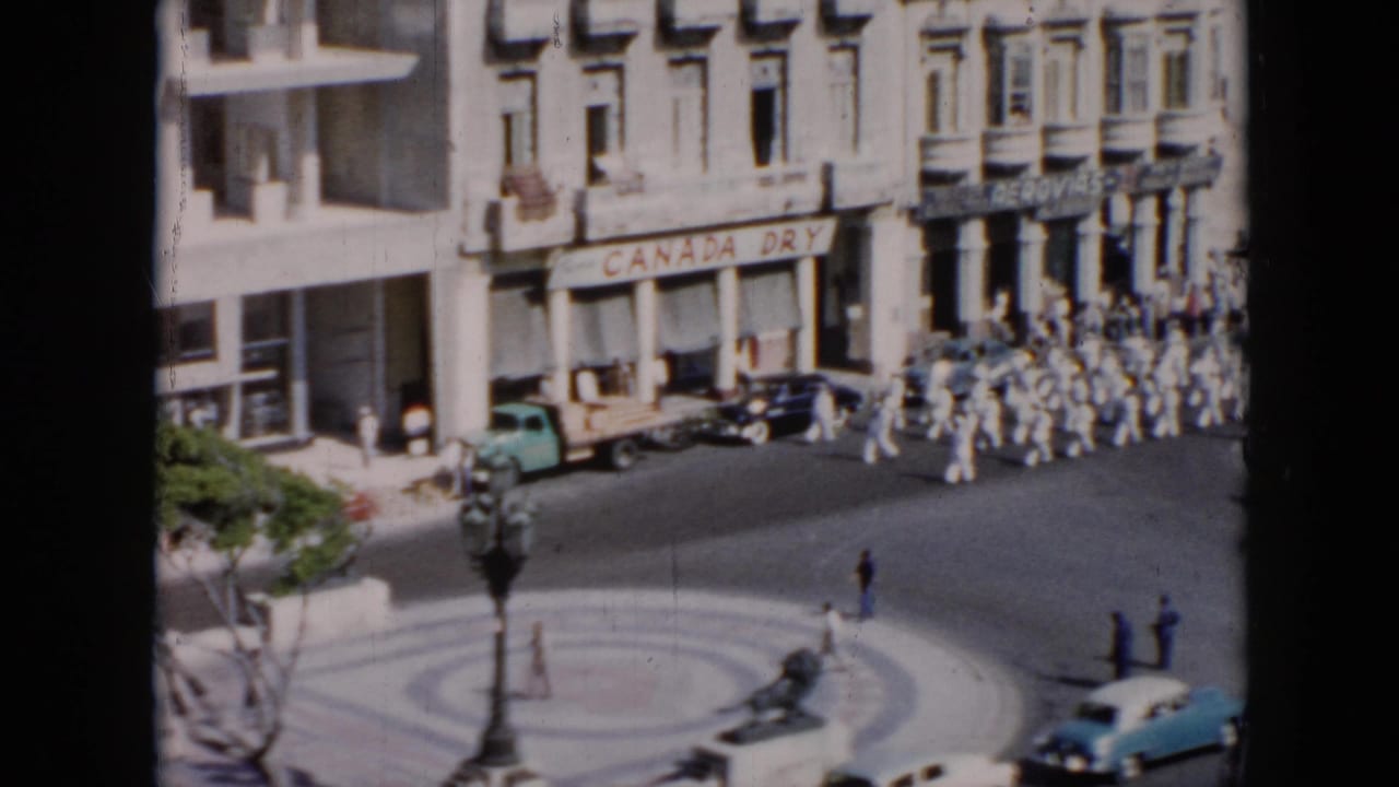 High-angle 1956 Cuba street view with white-uniformed men marching past storefronts, parked cars, pedestrians, and a circular plaza.