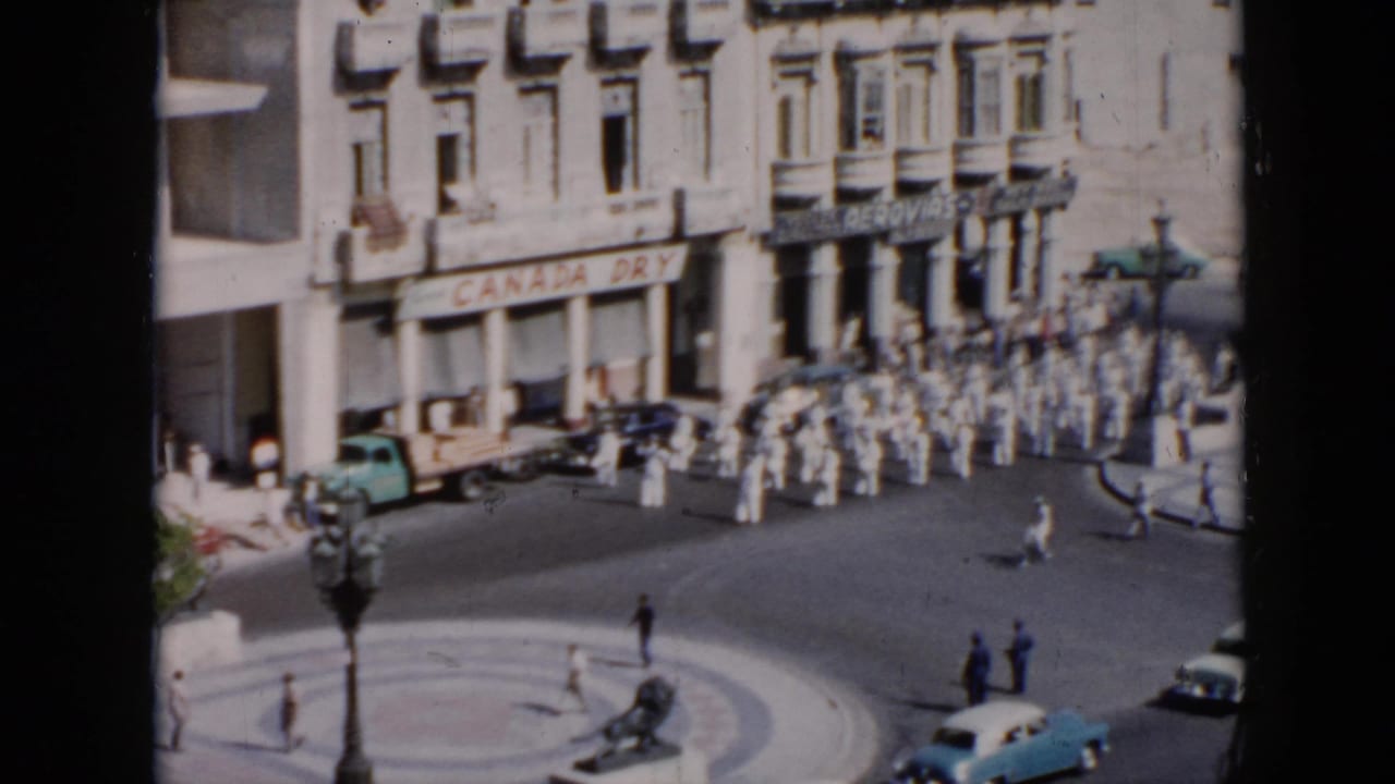 Marchers in white uniforms move across a sunlit street as cars and pedestrians remain nearby.