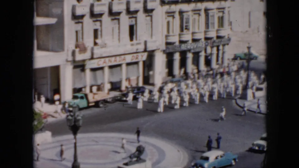 Elevated view of a 1956 Cuba street procession, with white-uniformed marchers passing storefronts, a light blue car, and pedestrians.