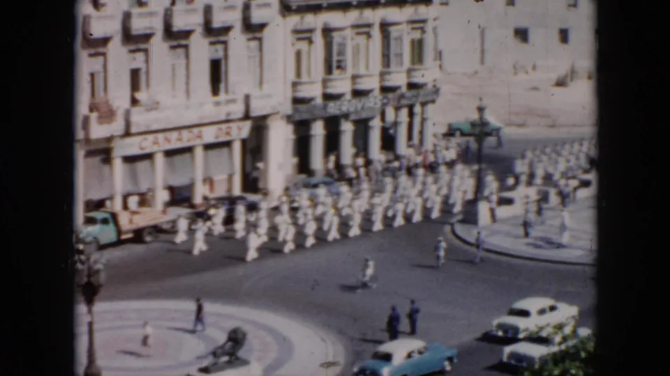 High-angle 1956 Cuba home-movie frame showing rows of white-uniformed men marching past a plaza, storefronts, cars, and pedestrians.