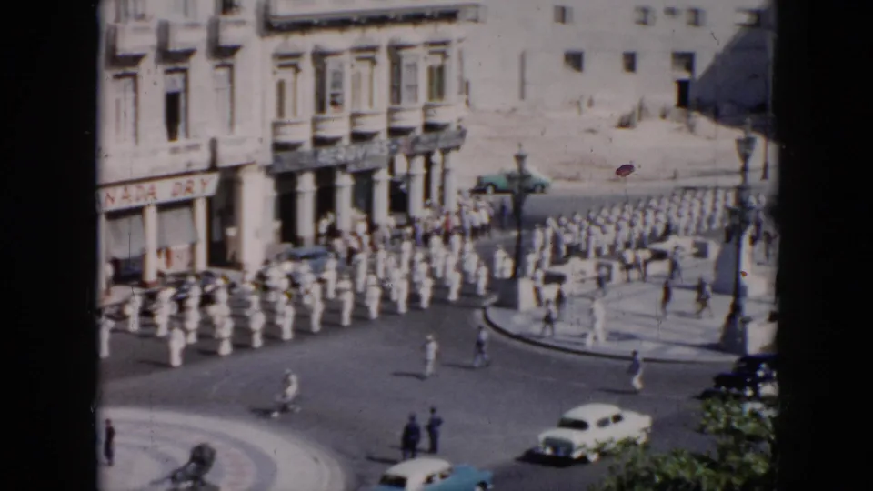 Wide elevated 1956 Cuba street scene with a long white-uniformed procession, circular plaza, pedestrians, mid-century cars, and storefront signage.
