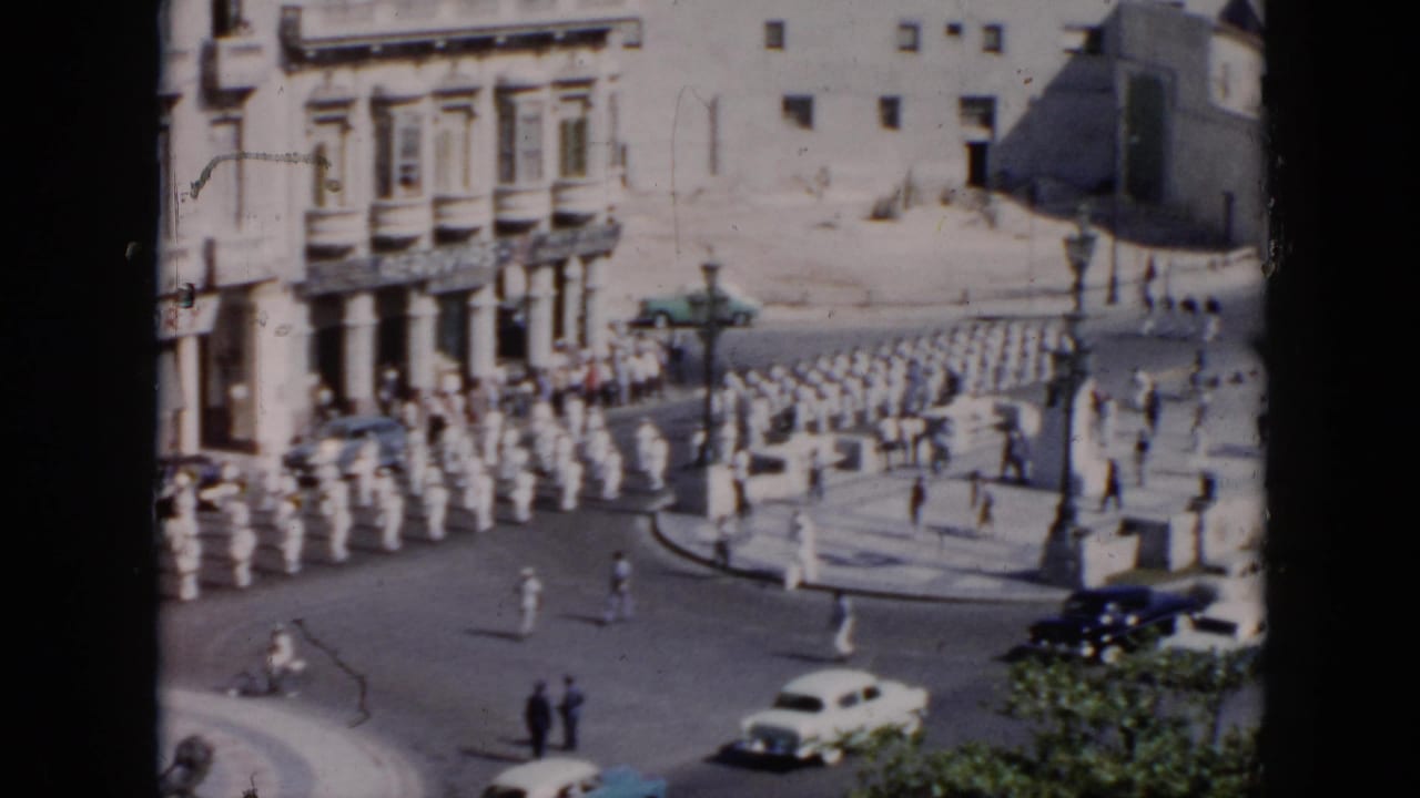 Soldiers in white uniforms march through a sunlit street near pale buildings and parked cars.