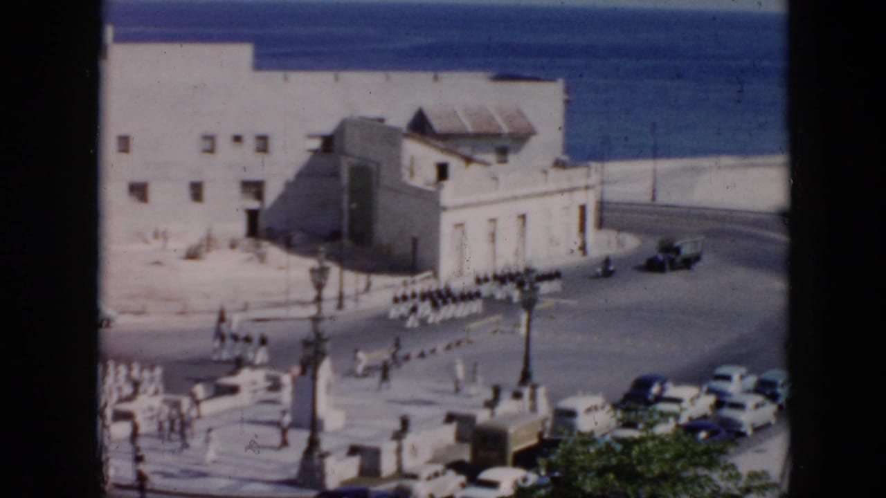 A column of white-uniformed soldiers marches along a coastal road with ocean visible beyond the buildings.