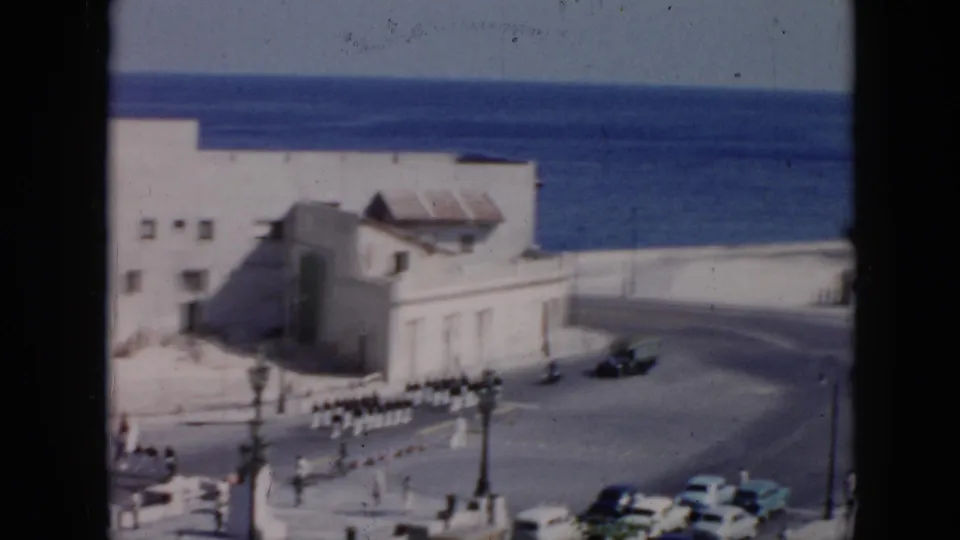 Elevated home-movie frame from 1956 Cuba showing soldiers in white uniforms marching near a seaside building, with mid-century cars, pedestrians, streetlamps, and ocean beyond.
