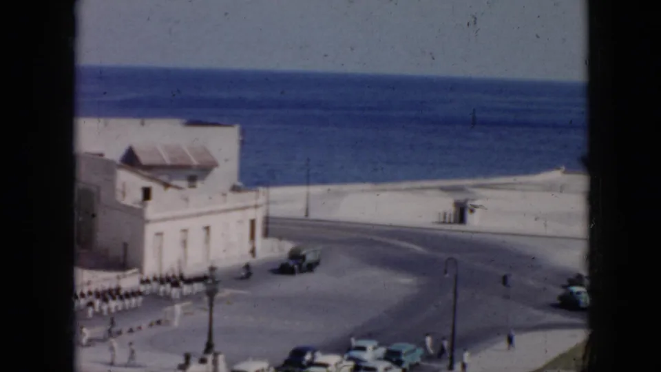 1956 Cuba home-movie view of a military parade crossing an oceanfront urban roadway, with white uniforms, pale buildings, street furniture, pedestrians, and classic cars visible.