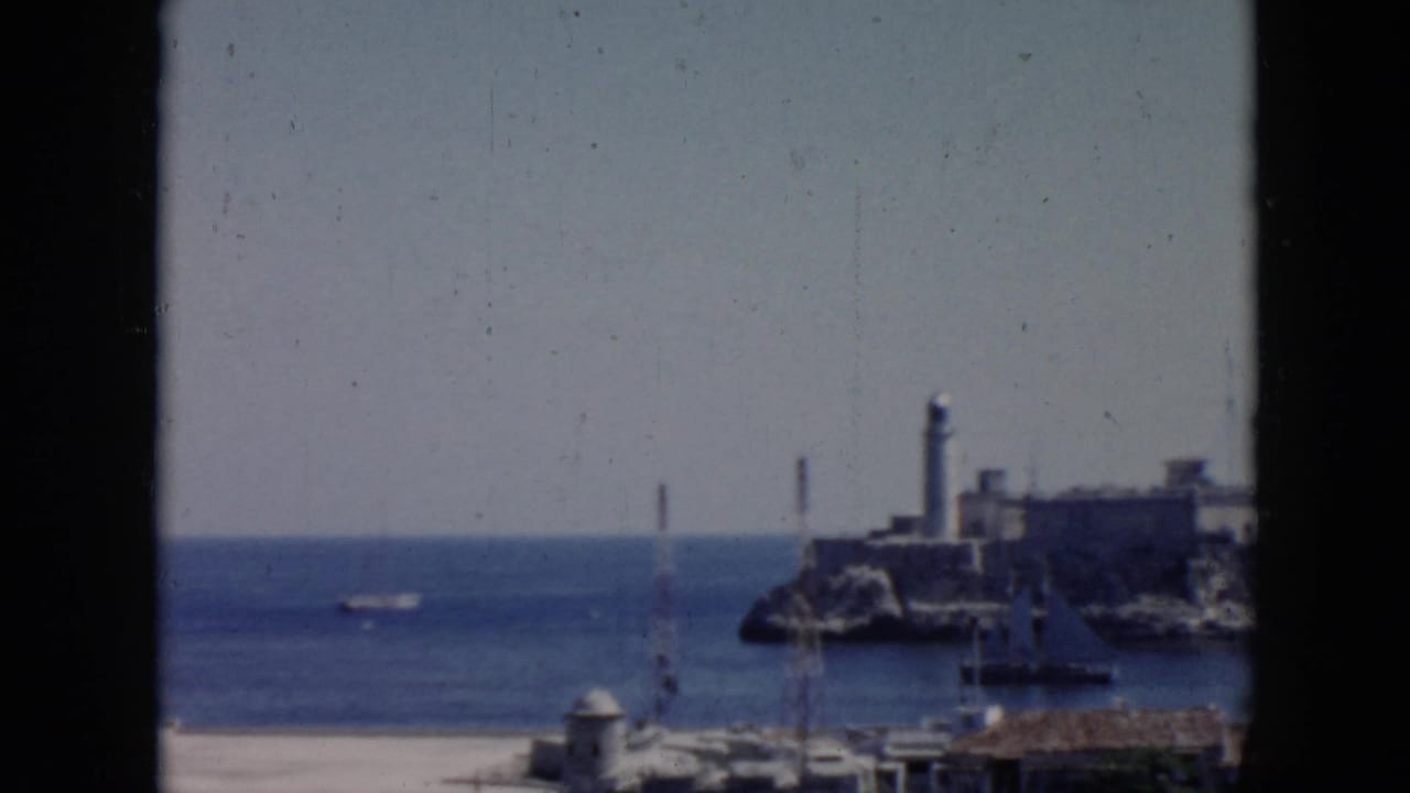 Similar harbor view with blue water, distant lighthouse, fortress walls, and a small boat offshore.