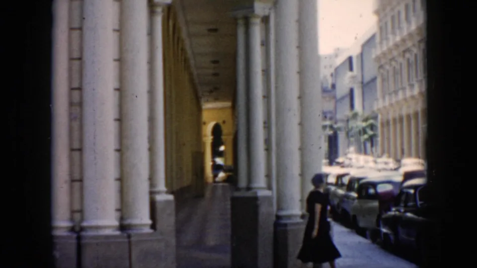 Havana street scene with a woman walking under tall columns beside rows of 1950s parked cars.