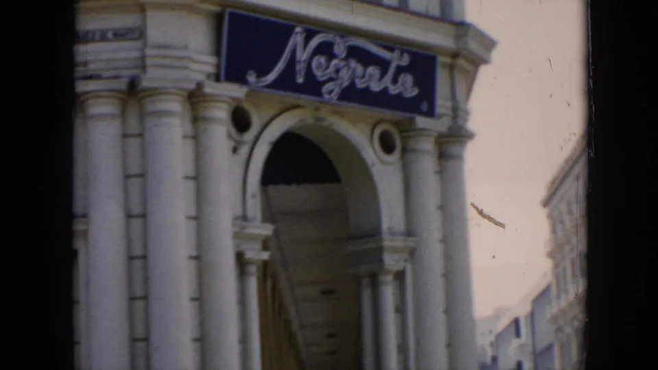 1956 Cuba frame with a prominent blue cursive sign over a columned archway and sunlit street facades in the background.