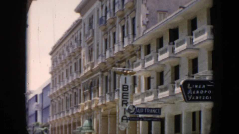 Cuba 1956 street footage frame with parked cars, palm trees, ornate balconies, and Iberia signage partly framed by dark film borders.