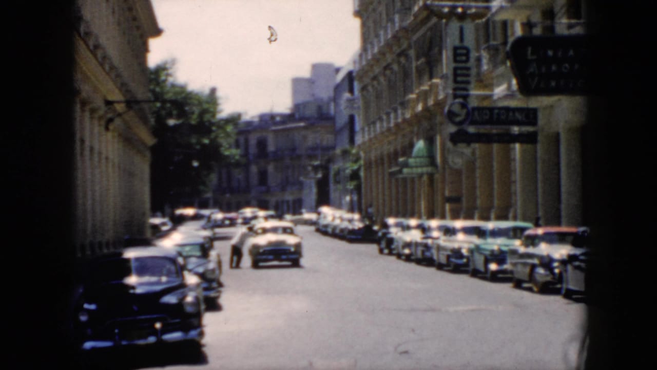 1956 Cuba street view with mid-century cars, columned facades, pedestrians, and a man beside a stopped car.