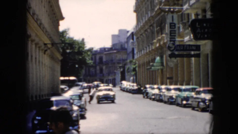 1956 Cuba home-movie frame showing a hat-wearing man in the foreground and a city street with cars behind him.