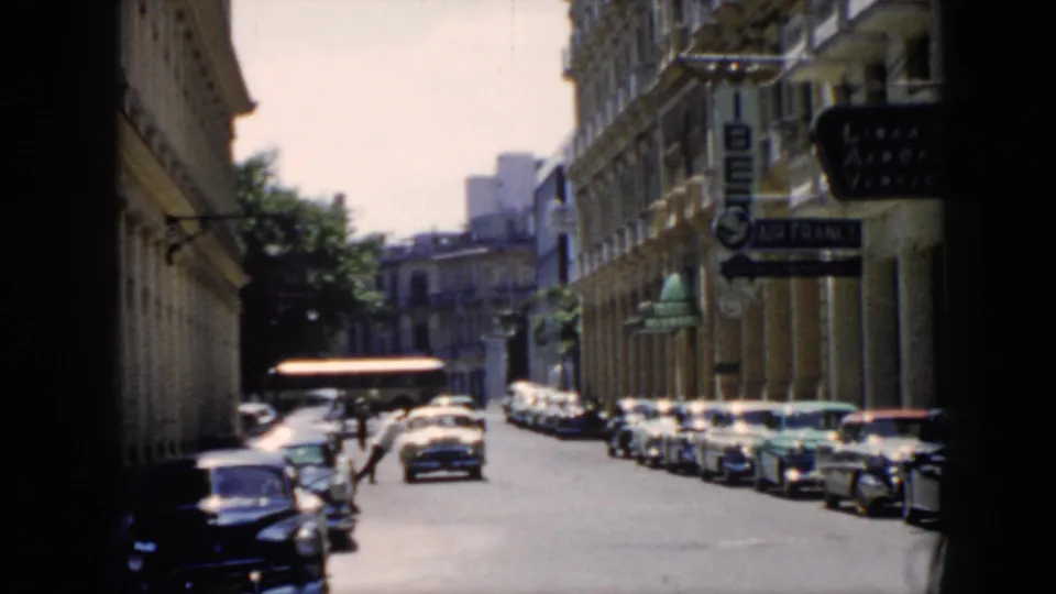 1956 Cuba street frame with a man leaning into a stopped car, parked automobiles, building signs, and pedestrians.