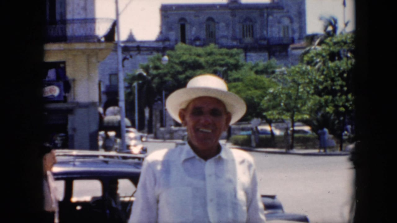 In supplied 1956 Cuba footage, a smiling man in a white hat and light shirt faces the camera on a street with older cars and balconies behind him.