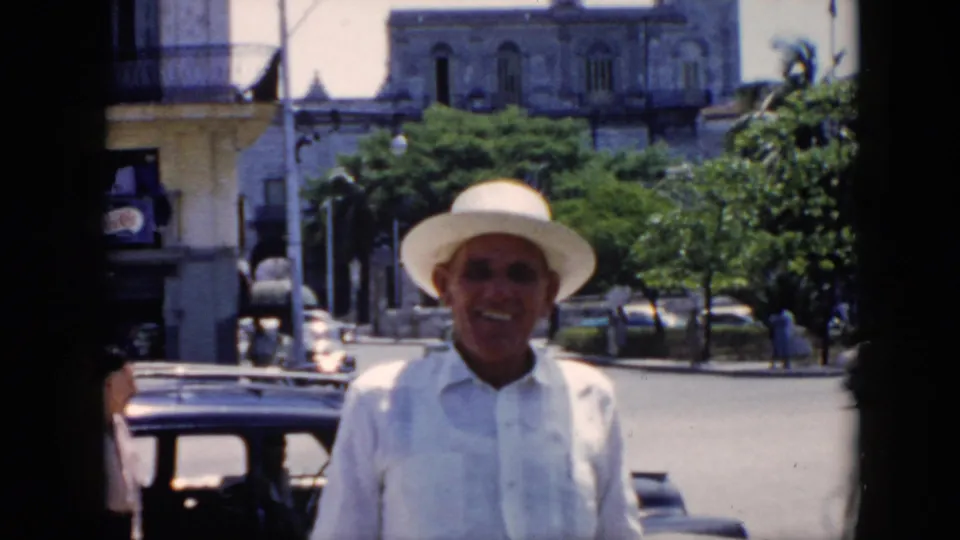 In supplied 1956 Cuba footage, a smiling man in a white hat and light shirt faces the camera on a street with older cars and balconies behind him.