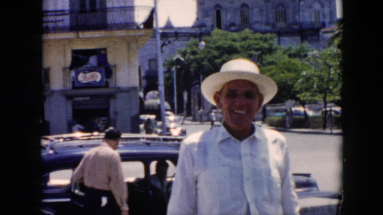 A smiling man in white clothing and hat stands near a dark mid-century car on a Cuban city street, with another adult visible by the vehicle.