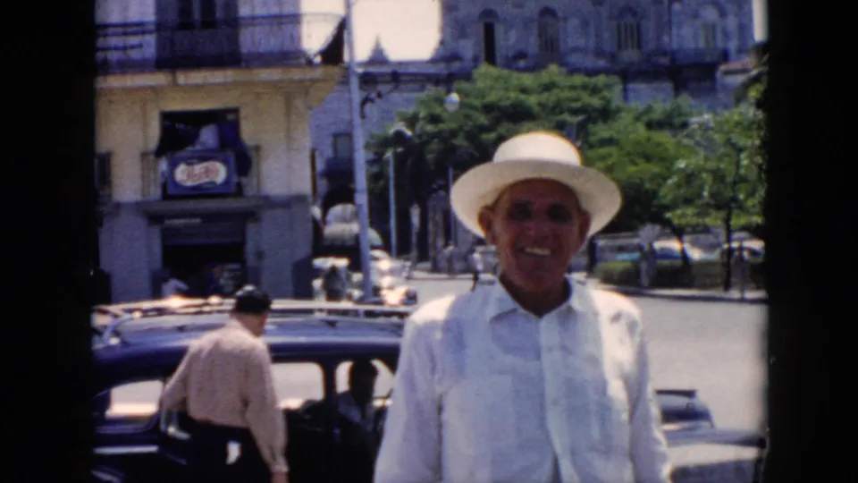 A smiling man in white clothing and hat stands near a dark mid-century car on a Cuban city street, with another adult visible by the vehicle.