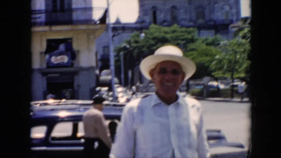 The man remains facing the camera in the supplied Cuba street footage, framed by an older car, a storefront sign, trees, and building balconies.