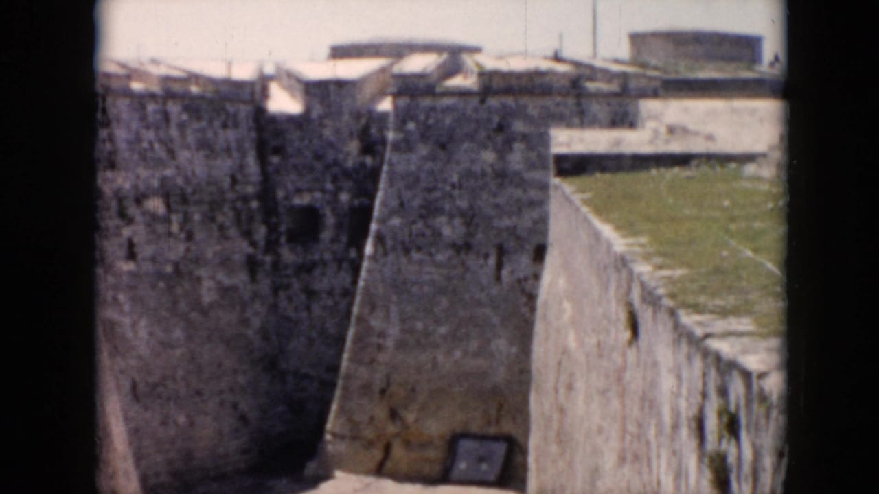 1956 Cuba home-movie frame showing rough stone fort walls, angled ramparts, grass above the wall, and a sunlit lower passage.
