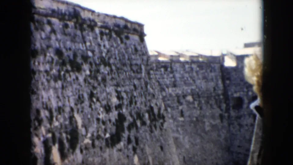 1956 Cuba frame showing an extended stone fort wall with dark patches, a sloped rampart, and pale daylight sky at the end of the shot.
