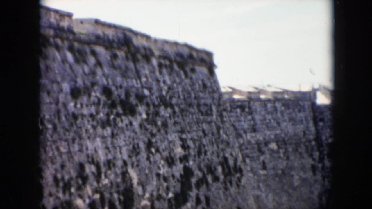 1956 Cuba home-movie frame looking up at a worn stone fortress wall from a shadowed opening, with pale sky above.