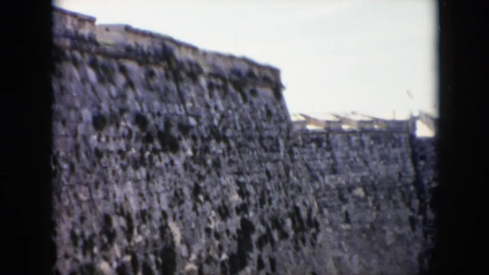 1956 Cuba home-movie frame looking up at a worn stone fortress wall from a shadowed opening, with pale sky above.