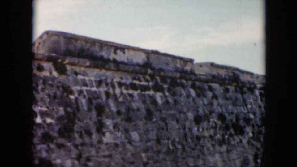 A low-angle 1956 Cuba home-movie view shows a broad weathered fortress wall and upper stone structure beneath a pale sky.