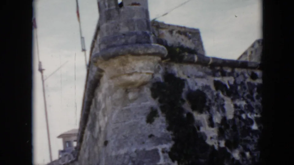 1956 Cuba 8mm frame showing the fortress corner, cylindrical turret, flagpoles, and a small distant building near the base.