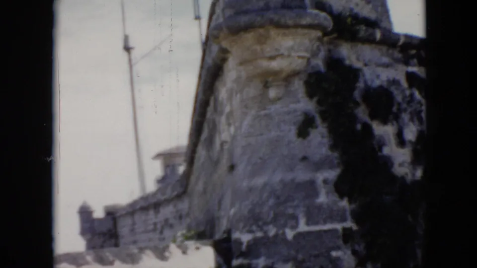 1956 Cuba 8mm frame with a moss-darkened fortress wall, near turret, flagpole, and smaller distant towers.