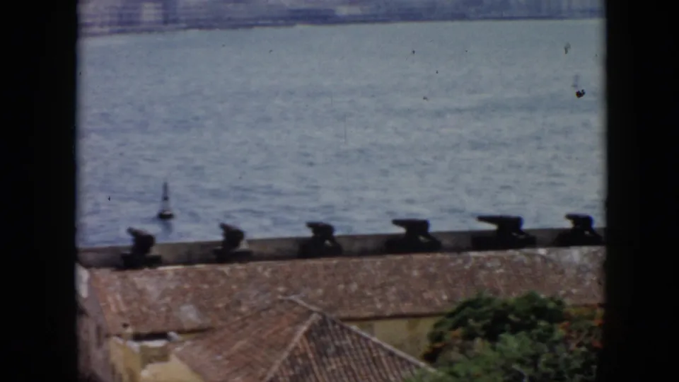 1956 Cuba 8mm view of cannon silhouettes along a waterfront roof with calm sea beyond.