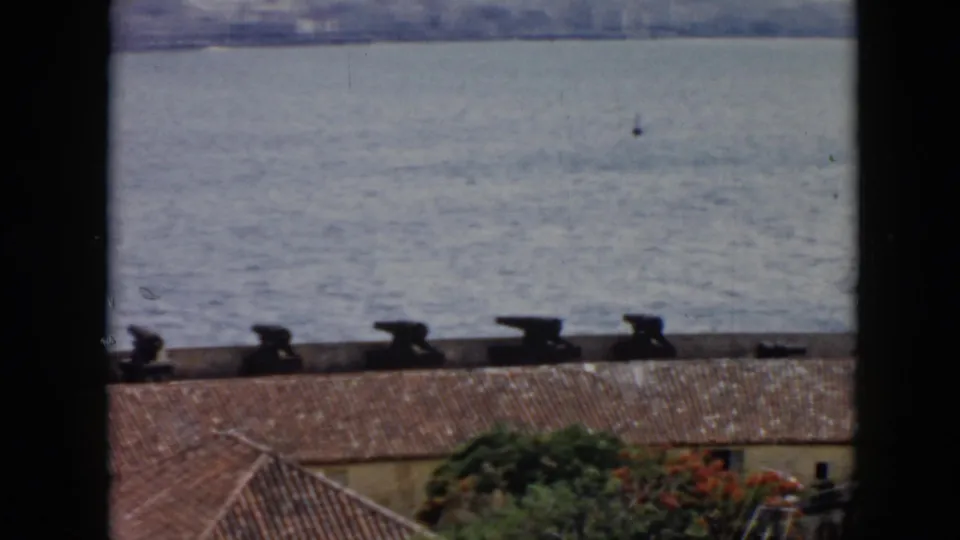 1956 Cuba home-movie frame with cannons, red roof tiles, greenery, and a small sailboat on the water.