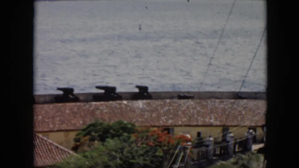 1956 Cuba 8mm frame showing cannons on a coastal fort edge above roof tiles, greenery, and open water.