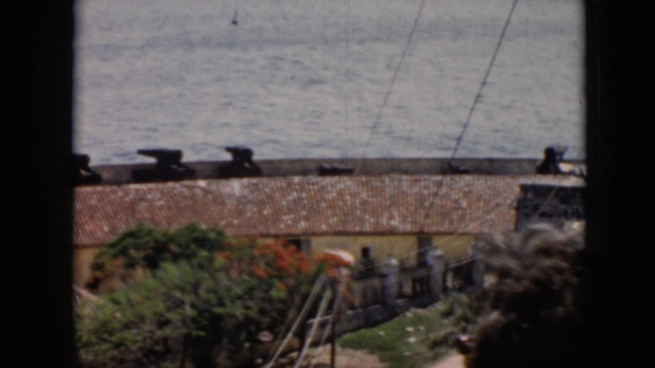 1956 Cuba home-movie frame looking through a dark window frame toward water, a terracotta roof, greenery, and a waterfront area.
