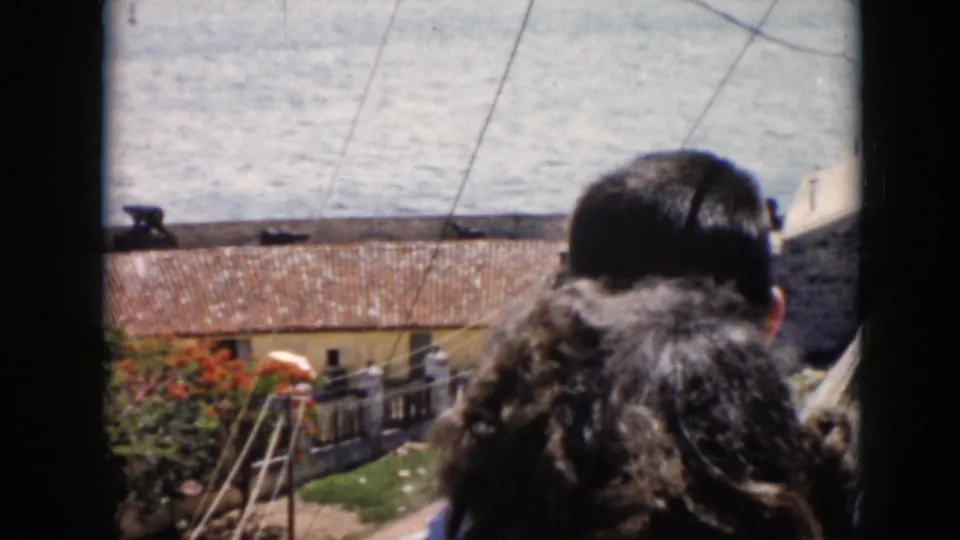 1956 Cuba home-movie frame where two people seen from behind partially block a coastal view through the window.