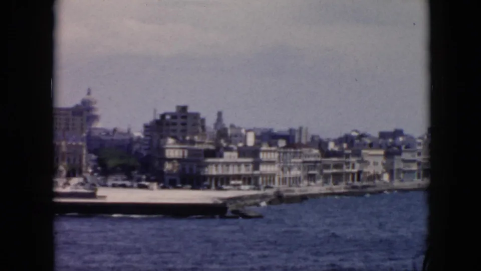 1956 Cuba coastal city view with dockside boats, trees, dense buildings, and choppy water from a moving boat.