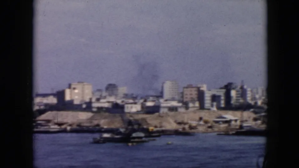 1956 Cuba harbor view with a lower shoreline, scattered buildings, cranes or dock equipment, and a small vessel on the water.