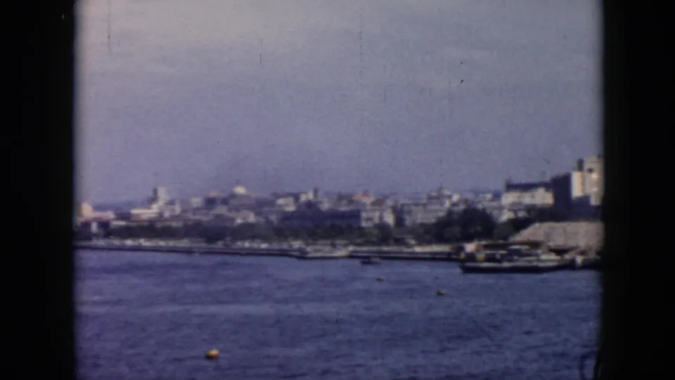 Grainy 1956 Cuba waterfront frame with calm water, distant city buildings, and small craft along the shore.