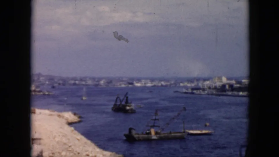 Wide 1956 Cuba harbor view showing industrial cranes, work boats, open water, and a distant coastal city skyline.