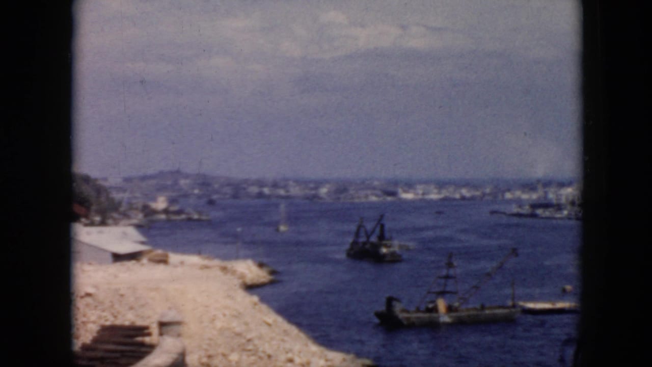 1956 Cuba home-movie frame showing a rocky harbor edge, small craft, and a distant waterfront under a hazy sky.
