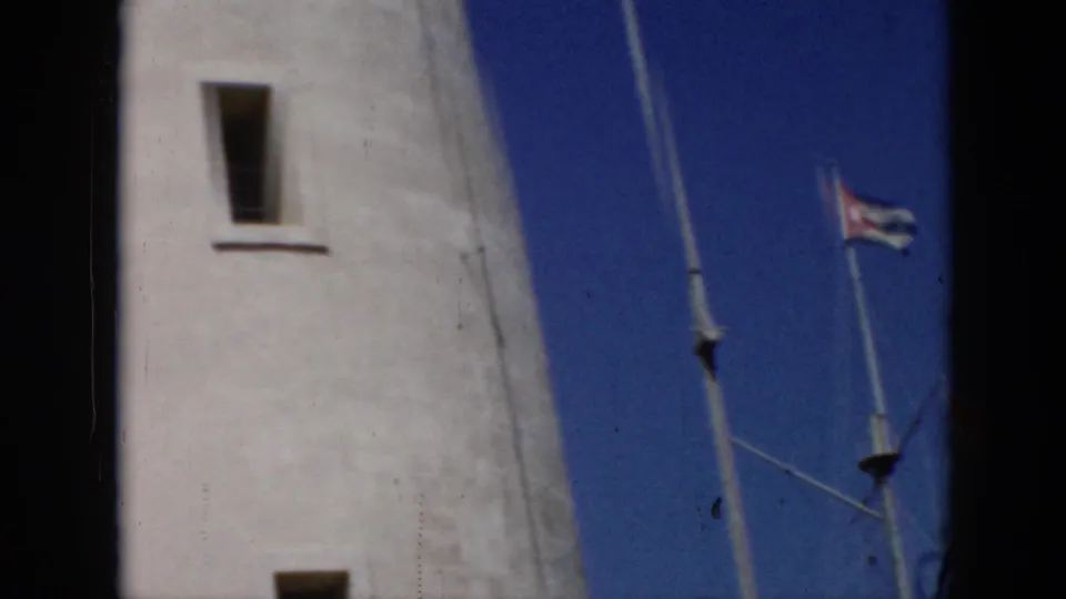 1956 Cuba 8mm frame showing a tall white lighthouse facade, narrow windows, blue sky, flagpole, and small Cuban flag.