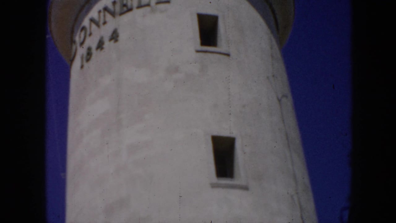1956 Cuba 8mm frame showing a low-angle view of a white lighthouse tower with partial DONNELL 1844 inscription against blue sky.