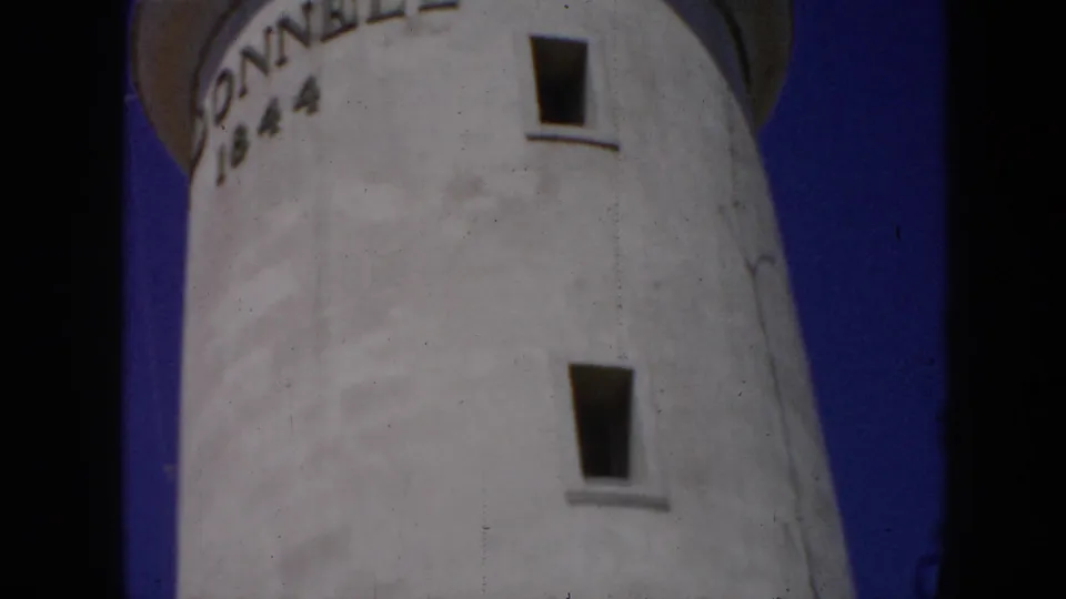 1956 Cuba 8mm frame showing a low-angle view of a white lighthouse tower with partial DONNELL 1844 inscription against blue sky.