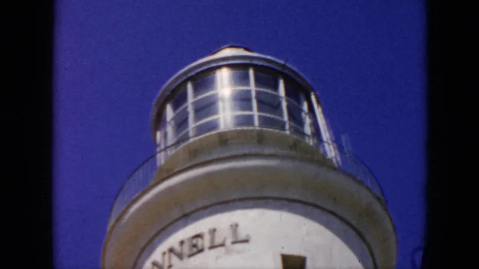 1956 Cuba 8mm frame showing the lighthouse top with glass lantern room, circular railing, blue sky, and partial tower lettering.