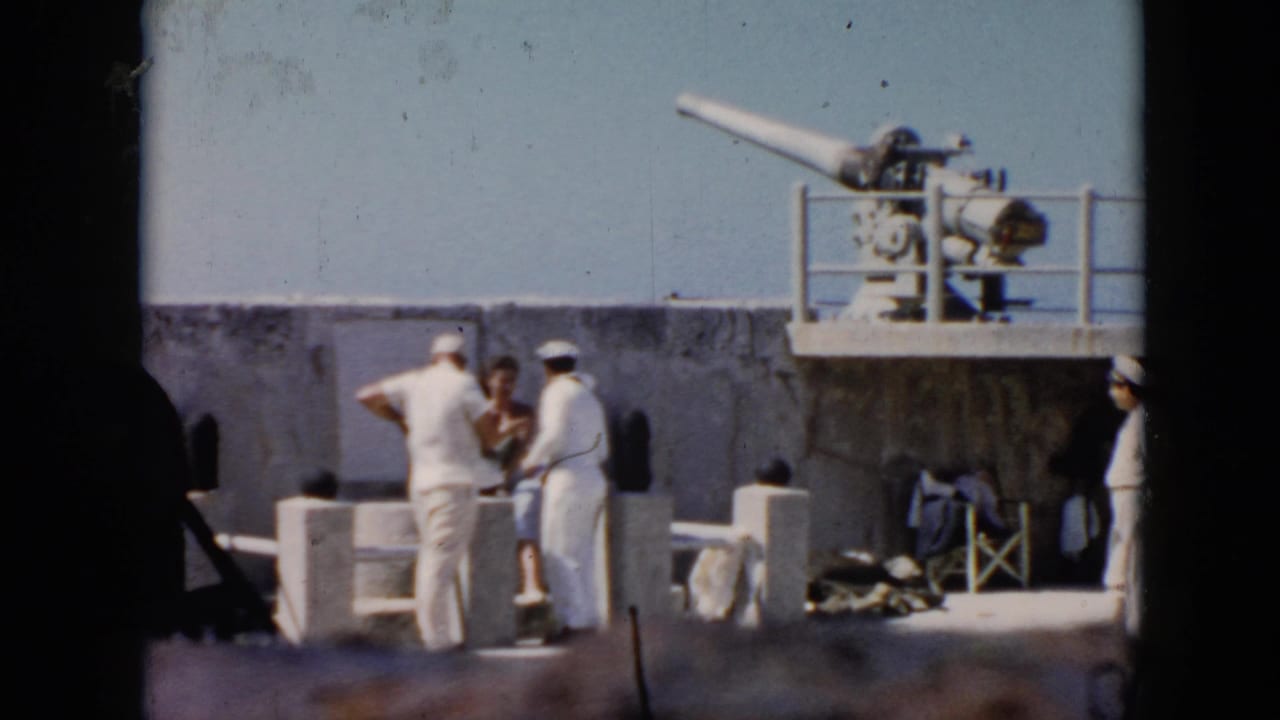 In Cuba in 1956, sailors in white uniforms and civilian visitors stand on a warship deck below a mounted naval gun.