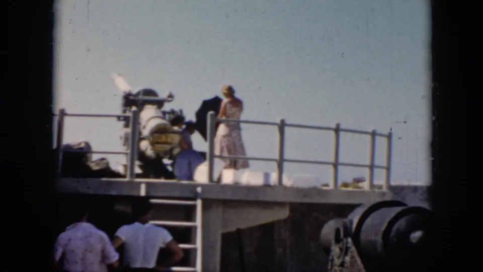 A woman in a light dress stands near a large naval gun on a Cuba warship deck in 1956, with other visitors below.