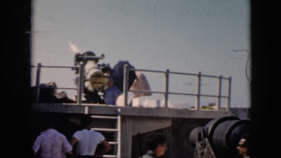 On a 1956 Cuba warship deck, a civilian visitor bends near the mounted naval gun while other people remain on the lower deck.